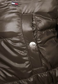 Mörk olivgrön puffjacka med detaljer som glänsande textur, sydda sektioner och en silverfärgad knapp med "THD." Inkluderar logotypaccenter ovanför.