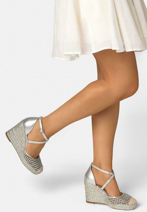 Piernas con alpargatas de cuña plateadas con tiras en el tobillo y malla decorativa, combinadas con un vestido blanco de vuelo.