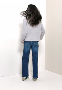 Chemise grise à manches longues associée à un jean bleu à jambes larges. Le jean présente des poches arrière avec des détails de couture. La tenue est complétée par des baskets grises et vertes.