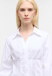 Camicia bianca a maniche lunghe con colletto, caratterizzata da cuciture verticali, una silhouette aderente e una texture di tessuto leggera e liscia.