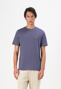 Joven vistiendo una camiseta de punto morada de manga corta con un pequeño logo de un pájaro amarillo en el pecho, combinada con pantalones beige.