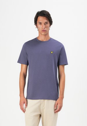 Jeune homme portant un t-shirt violet à col rond à manches courtes avec un petit logo d'oiseau jaune sur la poitrine, associé à un pantalon beige.