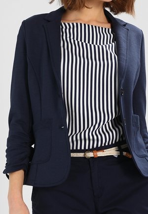 Kvinde iført en navy blazer over en navy og hvid vertikalt striped top, navy bukser og et vævet beige bælte med læderdetaljer.