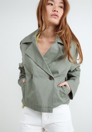Junge Frau mit langen Haaren, die eine hellgrüne, doppelreihige Jacke und weiße, zerrissene Jeans trägt, Hände in den Taschen, schaut zur Seite.