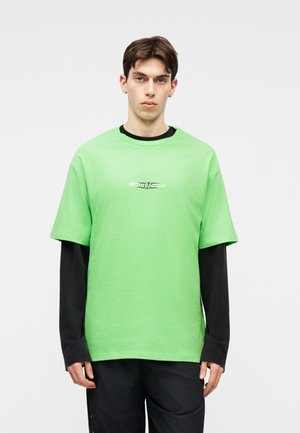 Ung mann med kort mørkt hår iført en lys grønn kortermet T-skjorte over en langermet svart skjorte, stående foran en enkel lys bakgrunn.