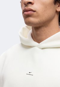 Νεαρό άτομο που φοράει λευκό hoodie με μικρό μαύρο λογότυπο Nike και κείμενο "NIKE STANDARD ISSUE" στο στήθος.