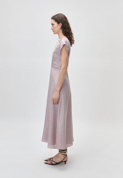 Γυναίκα με μωβ ανοιχτόχρωμο midi φόρεμα με κοντά μανίκια και μαύρα σανδάλια με λεπτά λουριά και χαμηλό τακούνι, σε προφίλ μπροστά σε απλό φόντο.