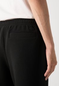 Pantalons de sport noirs en tissu texturé, avec une taille élastique et une poche arrière arborant un petit logo Lacoste. Main posée sur la hanche.