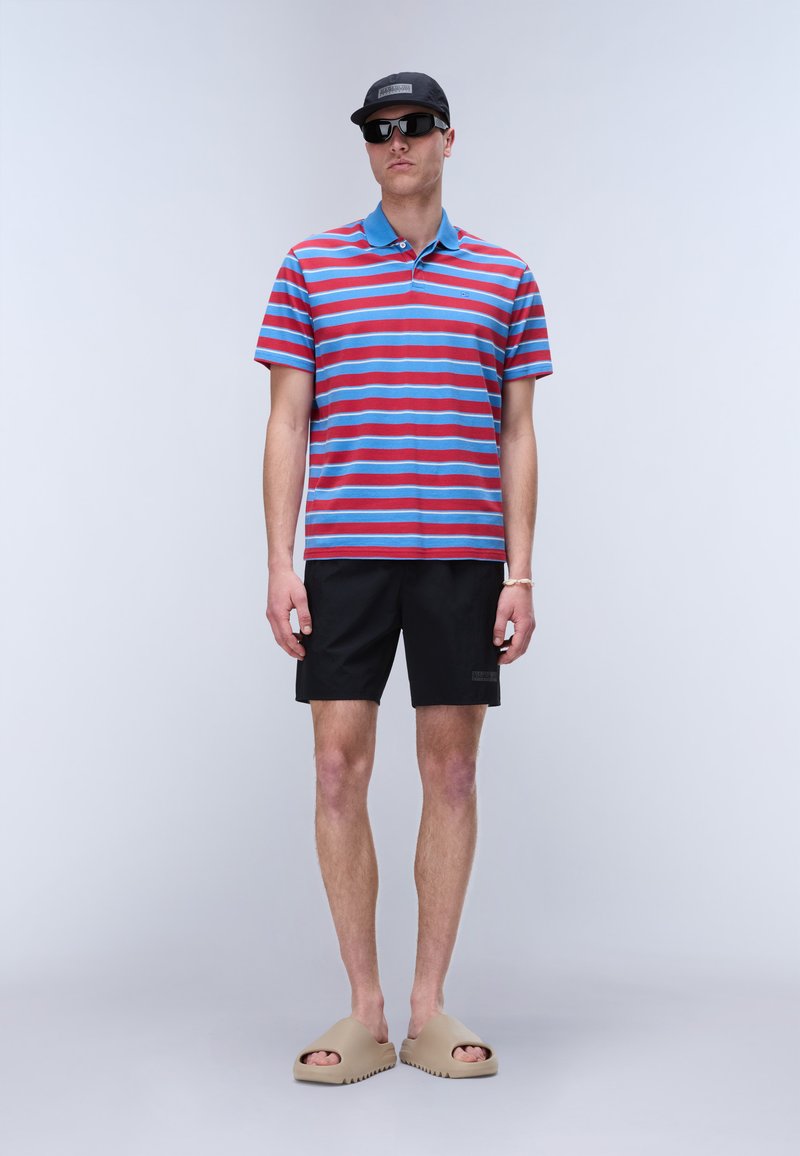 Mann trägt schwarze Kappe, schwarze Sonnenbrille, blau-rot gestreiftes Poloshirt, schwarze Shorts, beige Sandalen, steht vor grauem Hintergrund.