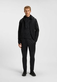 Completo sportivo nero composto da una giacca con cappuccio e cerniera, una maglietta a maniche lunghe con motivo e pantaloni aderenti con dettagli testurizzati e tasche.