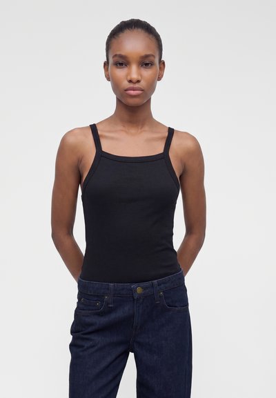 Jeune femme à la peau foncée et aux cheveux attachés porte un haut noir sans manches et un jean bleu foncé, debout devant un fond blanc uni.