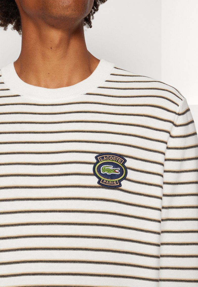 Wit gestreepte trui met horizontale zwarte en bruine strepen, geribbelde halslijn en een geborduurd Lacoste-logo op de borst.