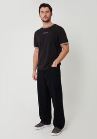 Hombre con barba vistiendo una camiseta negra con ribete beige, pantalones negros holgados y zapatillas grises, de pie contra un fondo claro y liso.