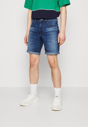 Shorts de denim en azul medio con un diseño desgastado, puños doblados, cinco bolsillos y una cintura estándar. Combinados con zapatillas blancas y calcetines.
