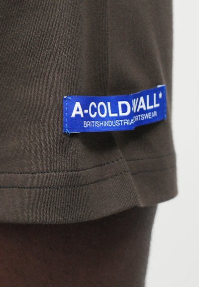 Hnedé bavlnené tričko s modrou tkanou nášivkou s nápisom "A-COLD WALL* BRITISH INDUSTRIAL SPORTSWEAR" blízko lemu, s textúrou rib. 