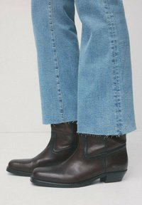 Ciemnobrązowe skórzane botki noszone z jasnoniebieskimi dżinsami z postrzępionymi nogawkami, stojące na jasnej podłodze.