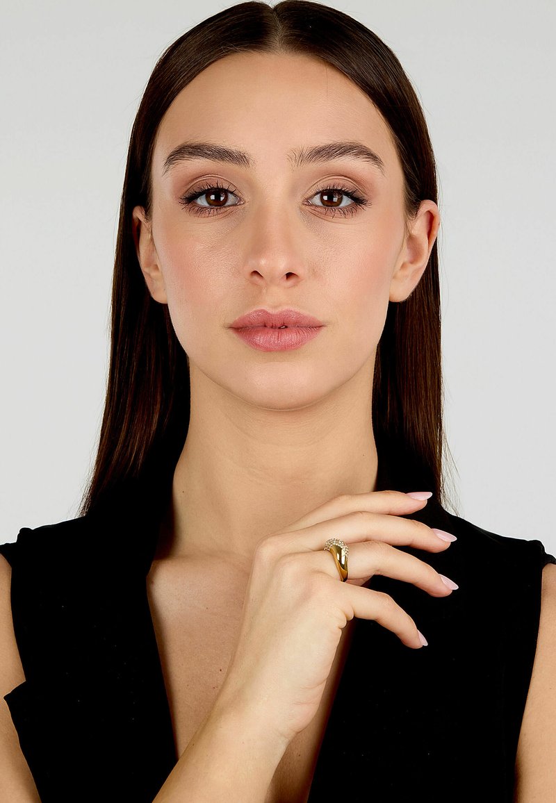 Anello d'oro con una finitura liscia, caratterizzato da un mazzo di piccole pietre su un lato. Il modello ha lunghi capelli marroni e un trucco neutro.