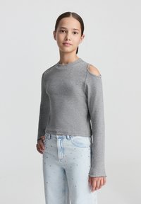 Maglione grigio a maniche lunghe con scollo alto e design spalle scoperte, abbinato a jeans azzurri chiari con decorazioni sparse.