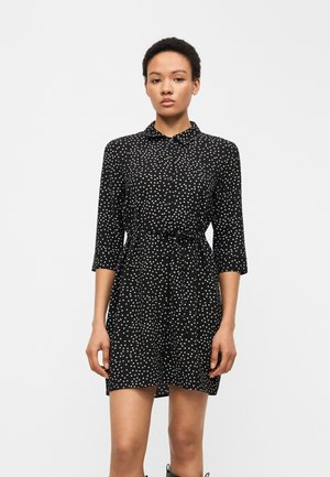 Kobieta w czarnej, zapinanej na guziki, krótkiej sukience w białe kropki, z rękawami o długości do łokcia i przewiązanym paskiem w talii, stoi na jednolitym tle.