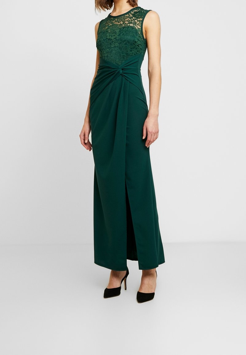 Robe vert foncé sans manches avec corsage en dentelle, détail twisté à la taille, fente à l'avant, portée avec des chaussures à talons noires sur fond uni.
