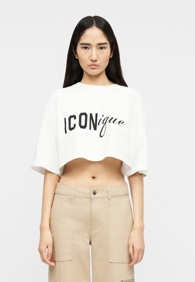 Jeune femme aux longs cheveux noirs portant un t-shirt blanc court avec le texte "ICONique" et un pantalon beige taille haute sur un fond uni.