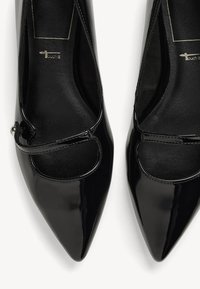 Tacones de punta afilada de charol negro con un acento de corte, que cuentan con una correa decorativa y una textura suave en la suela.