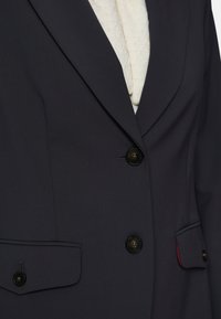 Navy blue tailored blazer made of smooth fabric, featuring two front pockets, black buttons, and a red accent on the pocket edge.