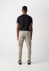 Bežové nohavice cargo so bočnými vreckami, slim fit, so zipsovými akcentmi, v kombinácii s čiernym tričkom a čiernymi teniskami, z pohľadu zozadu.