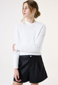 Jersey blanco esponjoso con cuello en V y mangas largas, combinado con pantalones cortos de imitación de cuero negro con bolsillos y detalles de botones en la parte frontal.