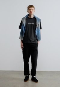 Μαύρο μπλουζάκι Calvin Klein με λευκό κείμενο, γκρι και μπλε φουλάρι σε στρώσεις, μαύρη φόρμα και μαύρα αθλητικά παπούτσια με καφέ σόλες.