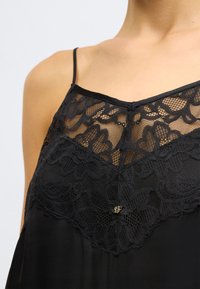 Camisolă neagră cu un panou din dantelă cu modele florale la decolteu, bretele subțiri reglabile și un mic accent decorativ.