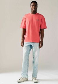 Koral oversized bomulds t-shirt med korte ærmer, der har et lille grafisk motiv på brystet, parret med lyseblå denim jeans og hvide sneakers.