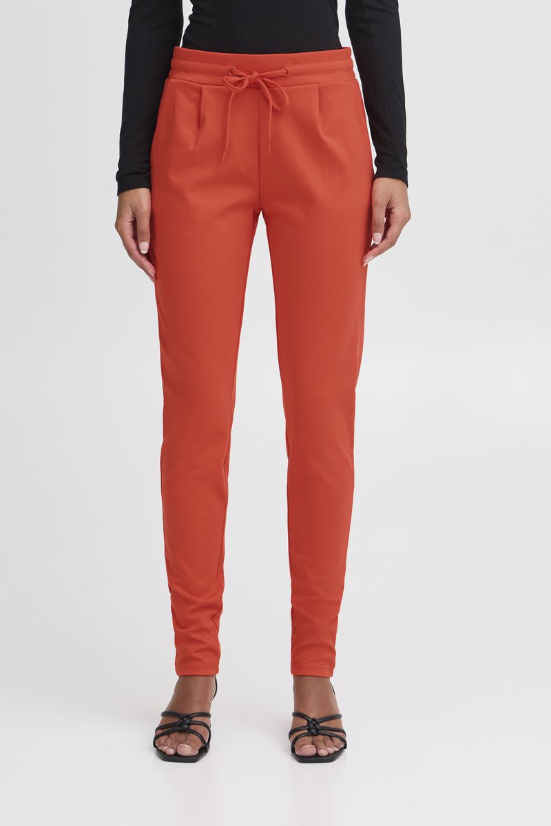 Woman wearing slim fit, high-waist, rust-colored pants with front drawstring, paired with black strappy sandals and a black long-sleeve top.