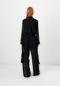 Crna prozirna košulja dugih rukava s ovratnikom, u kombinaciji s crnim hlačama širokog kroja koje imaju džepove i podesiv struk.