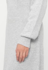 Una mano appoggiata contro un vestito a maglia grigio chiaro con motivi a coste e texture nella manica e nel corpo.