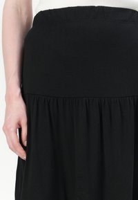 Femme portant une jupe noire taille haute froncée et un haut blanc, sa main gauche reposant à côté de la jupe.