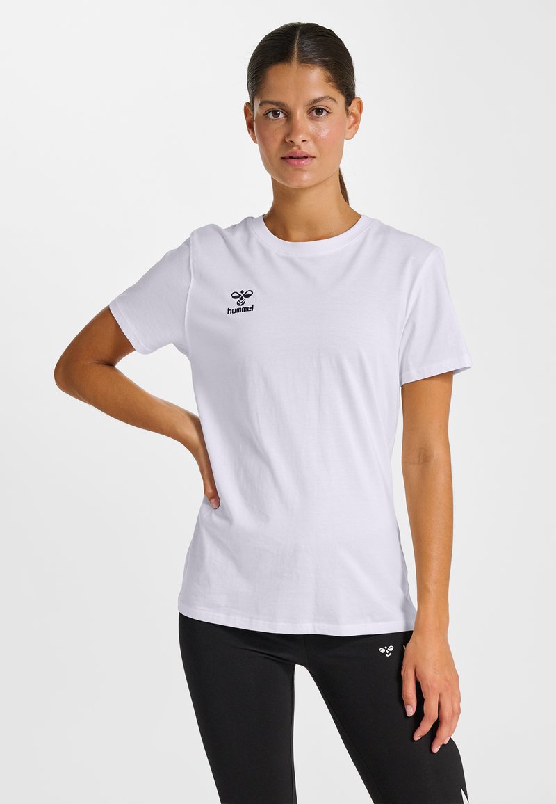 Femme aux cheveux noirs attachés en queue de cheval, portant un t-shirt blanc Hummel et un legging noir, debout avec une main sur la hanche, sur un fond blanc.