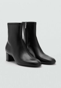 Botas de tobillo de cuero negro con cierre de cremallera, tacón cuadrado, textura suave y una silueta elegante. Presentan costura decorativa en la parte superior.