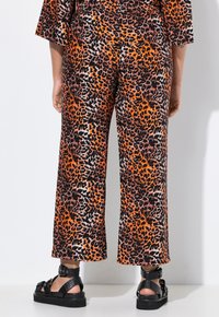 Leopardprint wijdlopende broek in oranje, zwart en crèmekleur, met een gladde textuur. Draagt zwart platform sandalen.