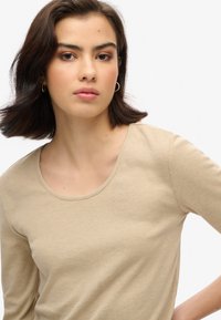Beżowa bluzka z długim rękawem i okrągłym dekoltem. Miękka, gładka tekstura, dopasowany krój z widocznymi szwami. Modelka nosi złote kolczyki w kształcie kół.