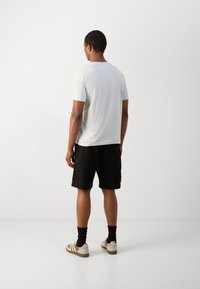 Lichtgrijs t-shirt met korte mouwen en een ronde hals, gecombineerd met zwarte shorts. Het model draagt gestreepte beige en roze sneakers.