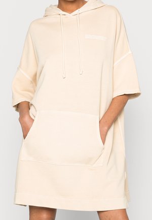 Robe à capuche beige oversize à manches courtes avec poche kangourou à l'avant et cordons, portée les mains dans les poches.