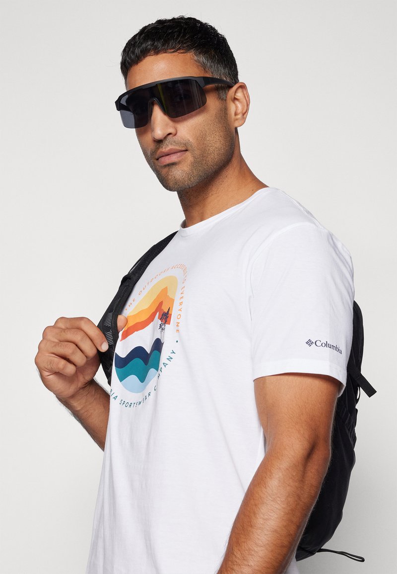 Camiseta blanca con un gráfico de montaña y ola multicolor. Gafas de sol oversized negras y una mochila negra con correas acolchadas.