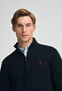 Suéter de punto negro con cuello con cremallera, que presenta un logotipo rojo en el lado izquierdo del pecho y está superpuesto a una camisa de cuello azul claro.