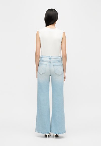 Γυναίκα με μακριά σκούρα μαλλιά, ντυμένη με λευκό αμάνικο μπλουζάκι και ανοιχτόχρωμο τζιν με φαρδιά πόδια, που στέκεται με την πλάτη προς τα εμπρός σε απλό φόντο.