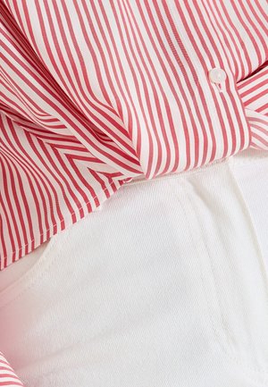 Chemise rayée rouge et blanche avec une fermeture à bouton, superposée sur un pantalon en denim blanc, mettant en valeur un tissu lisse et léger.