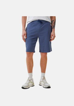 Mężczyzna w niebieskich szortach ze ściągaczem, białych trampkach i białych skarpetkach, stoi ze swobodnie opuszczonymi rękami na jednolitym tle.