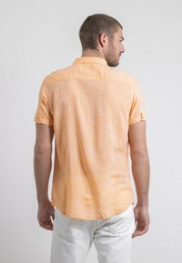 Chemise à manches courtes en orange clair, en tissu texturé avec un col boutonné, un ourlet droit et des manches retroussées. Étiquette visible sur le côté.