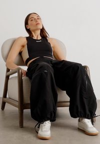 Pantaloni neri larghi con zip, abbinati a un top nero corto. Indossati con sneaker bianche e grigie, seduta su una sedia di colore chiaro.