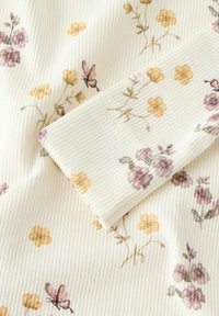 Prążkowana tkanina w kremowym kolorze z pastelowymi kwiatowymi wzorami w odcieniach żółtego i fioletowego. Posiada teksturowane wykończenie i detal złożonego rękawa.